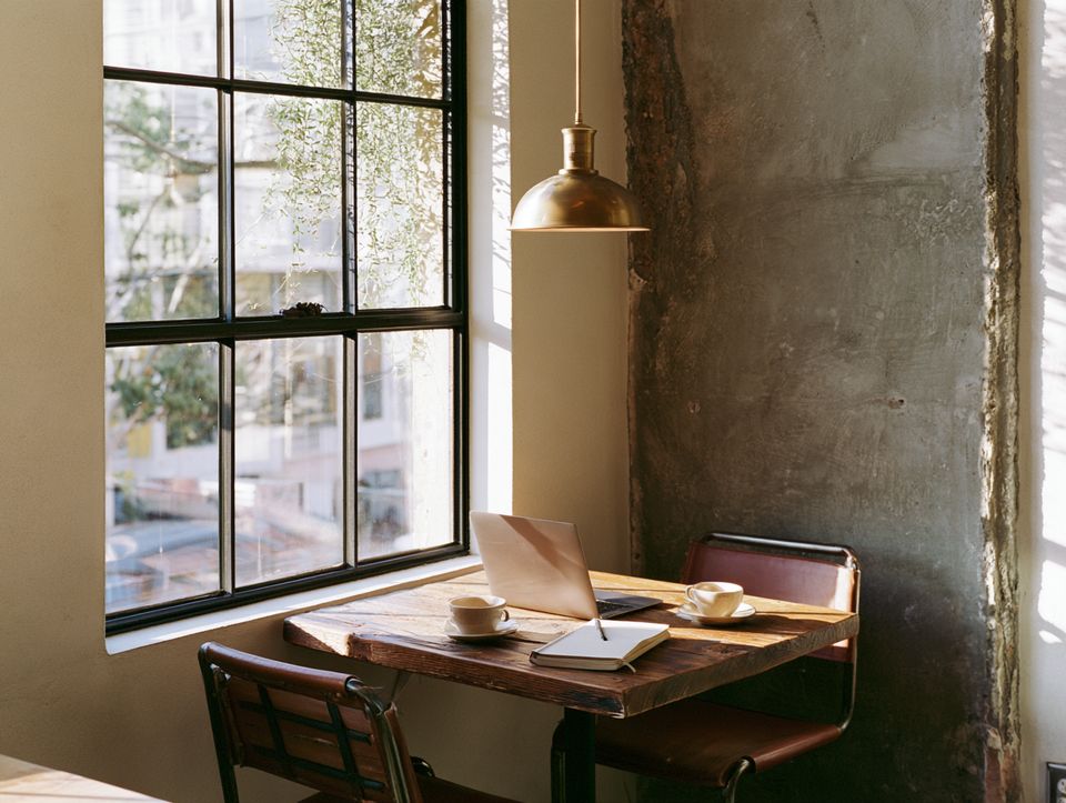 A quiet window table set for a conversation: two cups of coffee, a laptop and an open notebook under a brass lamp.